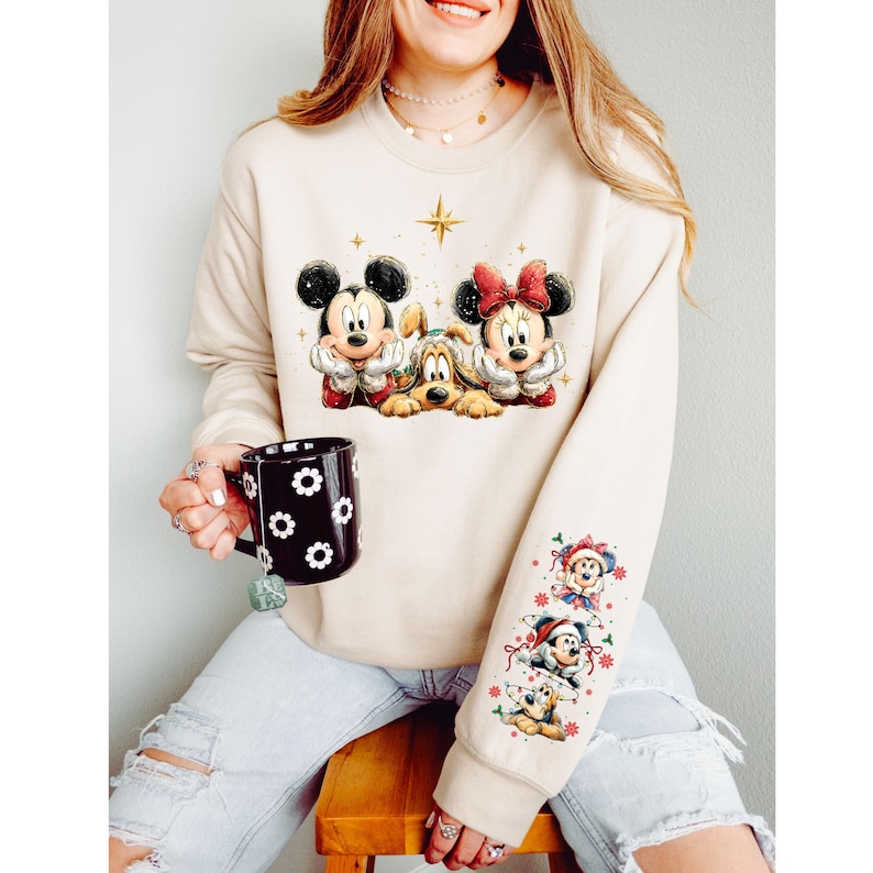 May include: Beige sweatshirt featuring Mickey Mouse, Minnie Mouse, and Pluto graphic. The sleeve has additional cartoon characters. A black mug with white flower designs is held in the hand.