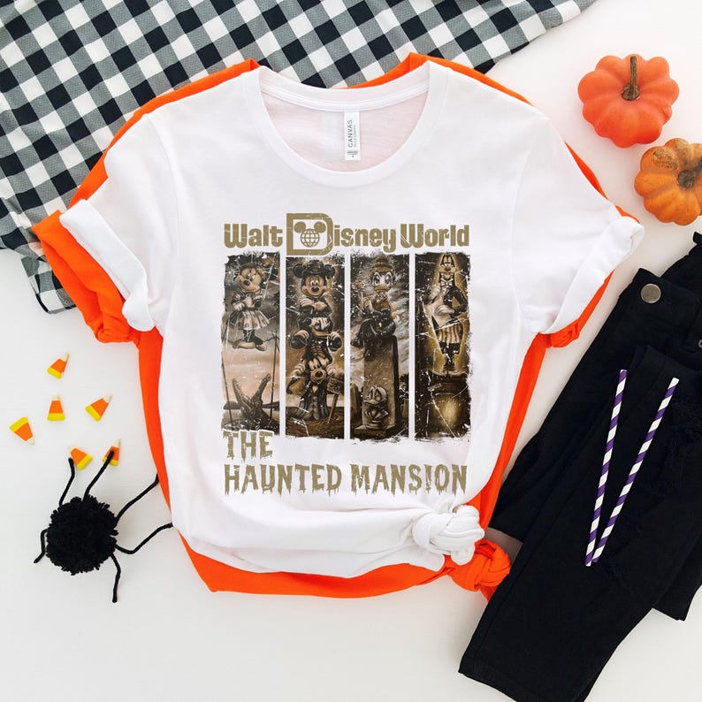 May include: A white t-shirt featuring a graphic of the Haunted Mansion attraction at Walt Disney World. The graphic depicts the iconic characters from the ride, including the Ghost Host, Constance Hatchaway, and the Hitchhiking Ghosts. The text "Walt Disney World" and "The Haunted Mansion" is printed in gold on the shirt.