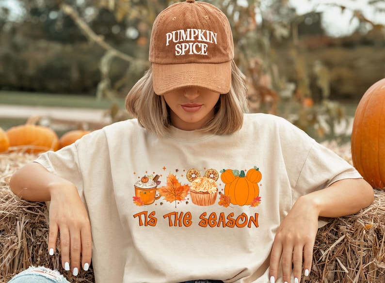 Pu&ograve; includere: T-shirt beige con grafica a tema autunnale e la scritta "Tis The Season". La grafica include un latte alla zucca speziato, foglie autunnali, un cupcake e zucche. Indossato un berretto da baseball di velluto a coste marrone con "PUMPKIN SPICE".