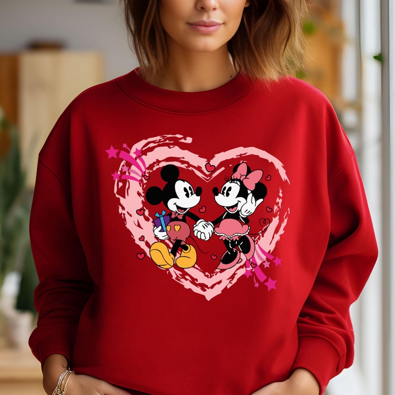 May include: A red sweatshirt featuring a graphic of Mickey and Minnie Mouse holding hands inside a pink heart. Mickey Mouse is holding a gift. Pink stars and hearts decorate the design. The sweatshirt is made of a soft material.
