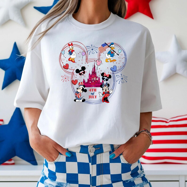 Peut inclure: T-shirt blanc avec un motif en forme de Mickey Mouse repr&eacute;sentant des personnages Disney c&eacute;l&eacute;brant le 4 juillet. Le motif comprend Donald Duck, Daisy Duck, Mickey Mouse et Minnie Mouse, avec des feux d'artifice et un ch&acirc;teau. Le texte "4th of July" est &eacute;galement inclus.