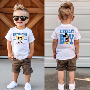 Puede incluir: Una camiseta blanca con un gr&aacute;fico de Mickey Mouse azul y amarillo y el texto "Birthday Boy" y "Est. 2018 Mickey Mouse". La camiseta la lleva un ni&ntilde;o peque&ntilde;o con el pelo casta&ntilde;o y gafas de sol. Tambi&eacute;n lleva pantalones cortos marrones y zapatillas blancas.