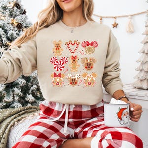 May include: A beige sweatshirt with a Christmas gingerbread themed design featuring Mickey Mouse and Minnie Mouse. The design includes gingerbread cookies shaped like Mickey Mouse and Minnie Mouse heads, gingerbread houses, candy canes, and red and white candy.