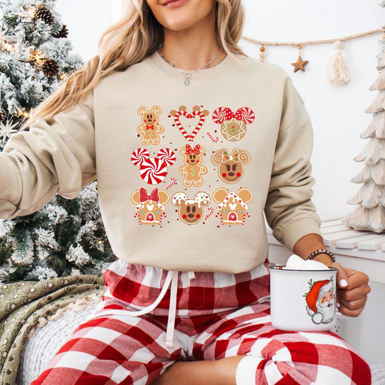 May include: A beige sweatshirt with a Christmas gingerbread themed design featuring Mickey Mouse and Minnie Mouse. The design includes gingerbread cookies shaped like Mickey Mouse and Minnie Mouse heads, gingerbread houses, candy canes, and red and white candy.