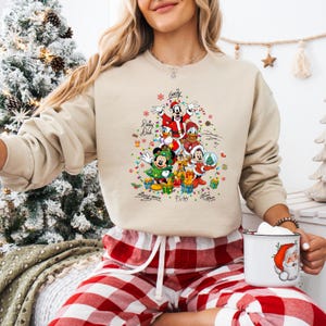 K&ouml;nnte beinhalten: Beiges Sweatshirt mit Rundhalsausschnitt und einem Weihnachtsbaumdesign mit Disney-Figuren wie Micky Maus, Goofy, Donald Duck und Pluto. Das Design enth&auml;lt farbenfrohe Akzente und die Figuren tragen festliche Kleidung. Das Sweatshirt wird mit einer rot-wei&szlig; karierten Pyjamahose kombiniert.