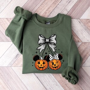 May include: An olive green sweatshirt featuring a Halloween-themed design. The design includes a large white bow, two jack-o'-lanterns with mouse ears, and a polka-dot bow. The pumpkins have smiling faces and are orange.