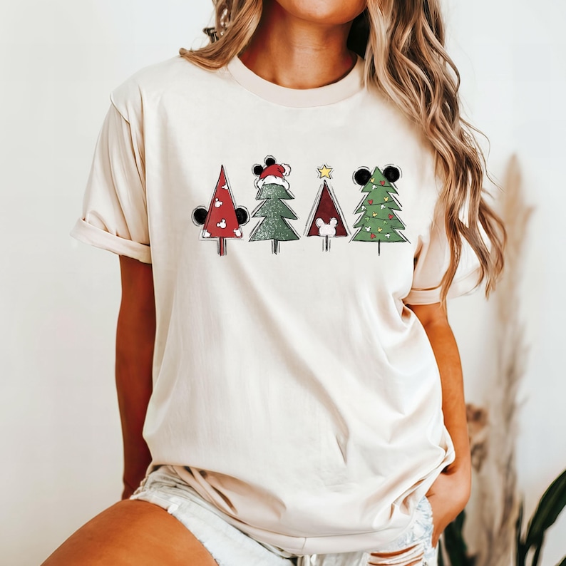 May include: A cream-colored t-shirt with a graphic of four Christmas trees decorated with Mickey Mouse ears. Each tree has a different design, including a red hat, a star, and ornaments.