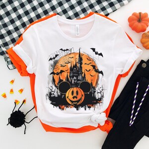 以下が含まれることがあります： 白いTシャツに、黒とオレンジのハロウィンデザインが施されています。ミッキーマウスの耳、カボチャの顔、満月を背景にしたシンデレラ城のシルエットが描かれています。