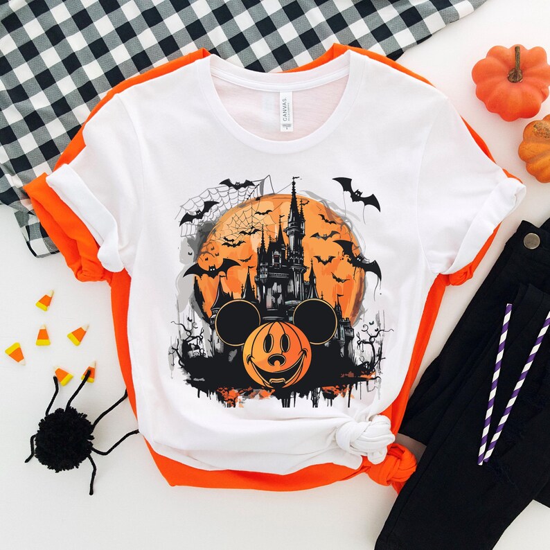 以下が含まれることがあります： 白いTシャツに、黒とオレンジのハロウィンデザインが施されています。ミッキーマウスの耳、カボチャの顔、満月を背景にしたシンデレラ城のシルエットが描かれています。