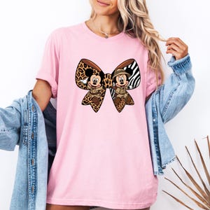 May include: Pale pink t-shirt with a large bow design featuring Mickey and Minnie Mouse in safari attire. The bow incorporates leopard print, zebra stripes, and brown detailing. The shirt is a light pink colour.