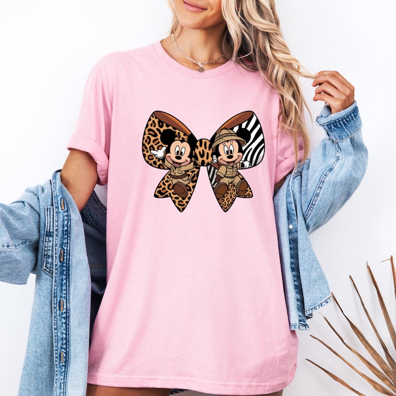 May include: Pale pink t-shirt with a large bow design featuring Mickey and Minnie Mouse in safari attire. The bow incorporates leopard print, zebra stripes, and brown detailing. The shirt is a light pink colour.