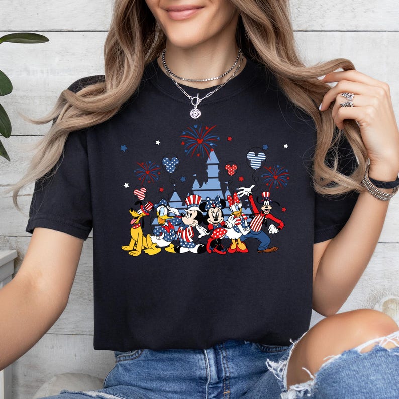 K&ouml;nnte beinhalten: Marineblaues T-Shirt mit einem Design von Disney-Figuren in patriotischer Kleidung, darunter Mickey und Minnie Mouse, Donald Duck und Goofy, mit Feuerwerk und Schlosssilhouetten. Das Design verwendet die Farben Rot, Wei&szlig; und Blau.