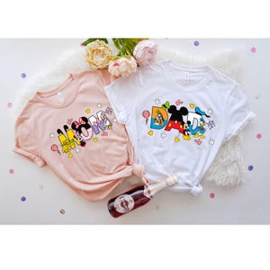 May include: Two t-shirts with a pink and white color scheme. The pink shirt has the word "MOM" spelled out with Disney characters. The white shirt has the word "DAD" spelled out with Disney characters.