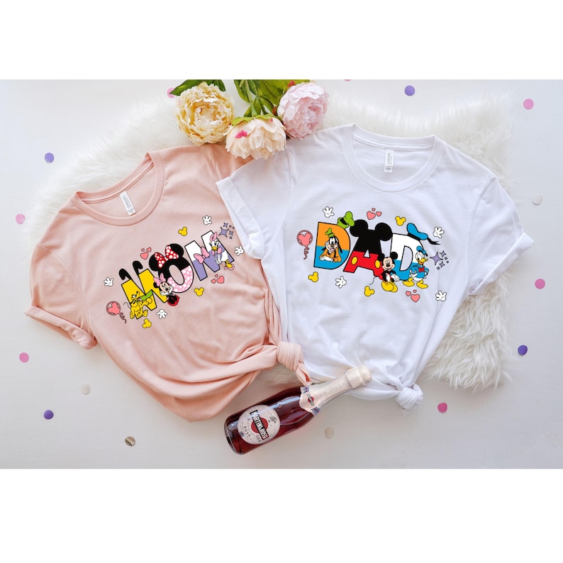 May include: Two t-shirts with a pink and white color scheme. The pink shirt has the word "MOM" spelled out with Disney characters. The white shirt has the word "DAD" spelled out with Disney characters.