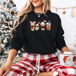 May include: Black sweatshirt featuring four festive coffee cup designs with holiday-themed toppings. The cups include Mickey Mouse ears, a Santa hat, and a shamrock. The person is wearing red and white plaid pajama pants and holding a Santa mug.