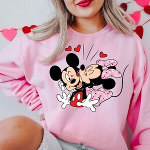 May include: Pink sweatshirt featuring a graphic of Mickey Mouse and Minnie Mouse kissing. The graphic is surrounded by small red hearts.
