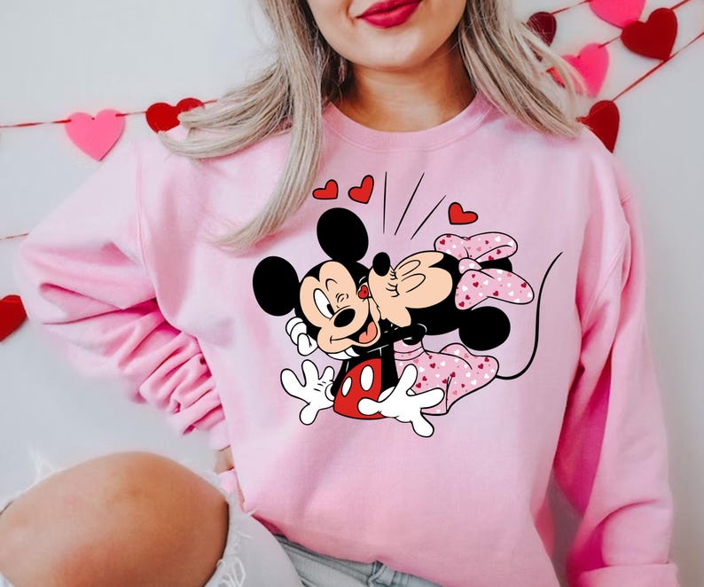 May include: Pink sweatshirt featuring a graphic of Mickey Mouse and Minnie Mouse kissing. The graphic is surrounded by small red hearts.