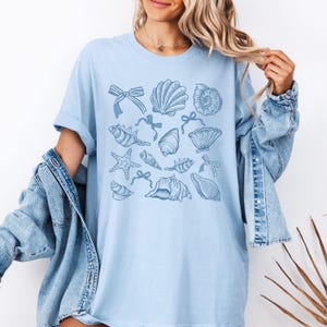 Pu&ograve; includere: Una maglietta blu chiaro con un motivo di conchiglie blu e stelle marine. Le conchiglie sono disposte in modo casuale e alcune hanno fiocchi.