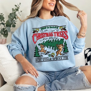 K&ouml;nnte beinhalten: Hellblauer Pullover mit einem Aufdruck von Chip und Chap, die einen Weihnachtsbaum tragen. Der Text lautet "Chip & Dale's Christmas Trees, free delivery, Pine-Cedar-Spruce-Fir."