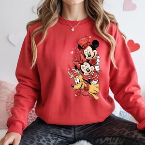 Puede incluir: Una sudadera roja con un gr&aacute;fico de Mickey y Minnie Mouse, y Pluto, con atuendos de San Valent&iacute;n. Mickey Mouse lleva un sombrero de copa, Minnie Mouse tiene un lazo y Pluto tiene alas de &aacute;ngel. Tambi&eacute;n se presentan corazones y rosas.