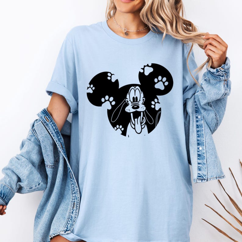 May include: Pale blue t-shirt with a black and white graphic of Pluto within a Mickey Mouse head shape. The Mickey Mouse head is filled with paw prints. The shirt has a relaxed fit.