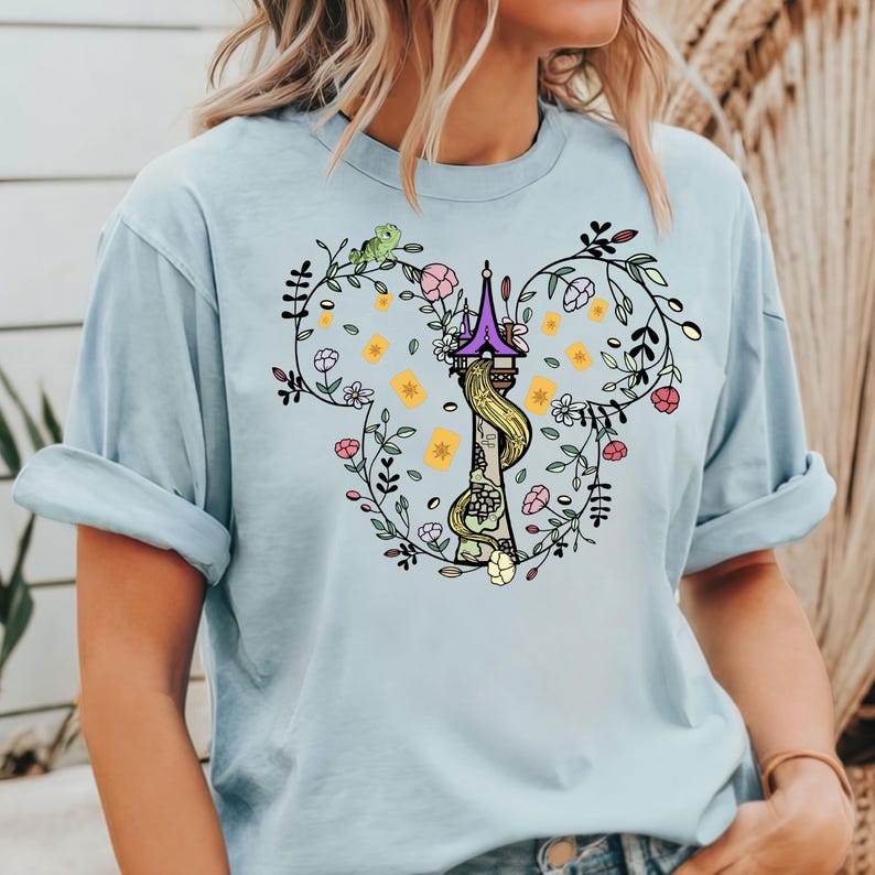 May include: A light blue t-shirt with a floral design featuring a tower and a long braid of hair. The design is in the shape of a Mickey Mouse head.