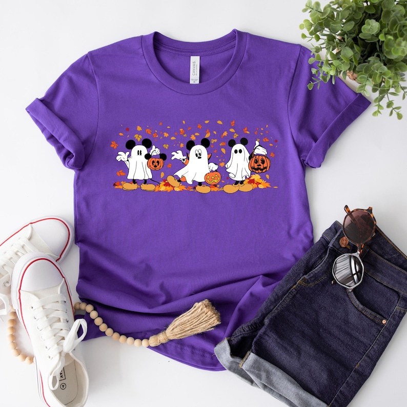 May include: Purple t-shirt with a graphic of three Mickey Mouse ghosts holding jack-o'-lanterns. The ghosts are wearing Mickey Mouse ears and are surrounded by falling leaves.