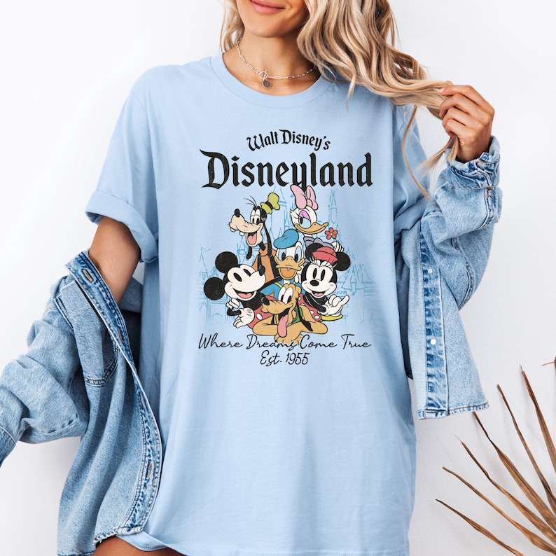 May include: Light blue t-shirt featuring the Disneyland logo and a graphic of Mickey Mouse, Minnie Mouse, Donald Duck, Goofy, and Pluto. The text "Where Dreams Come True Est. 1955" is also printed on the shirt.
