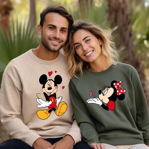 May include: Two sweatshirts, one tan and one olive green, featuring Mickey and Minnie Mouse. Mickey is on the tan sweatshirt with hearts, and Minnie is on the green sweatshirt blowing a kiss. Both sweatshirts have raglan sleeves.