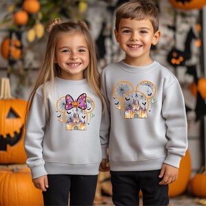 May include: Two children wearing matching gray sweatshirts with Halloween-themed designs. The sweatshirts feature a castle, spiderwebs, bats, and Mickey Mouse ears. One sweatshirt has a purple bow with a pumpkin. The other has a castle design.