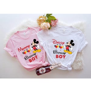 May include: Two t-shirts with a Mickey Mouse design. The pink shirt says "Mommy of the Birthday Boy" and the white shirt says "Daddy of the Birthday Boy".