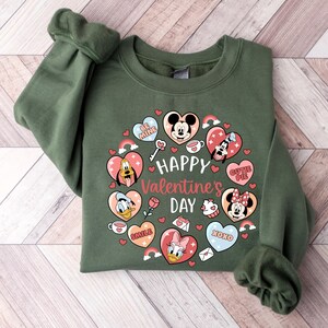 K&ouml;nnte beinhalten: Ein gr&uuml;nes Sweatshirt mit einem Valentinstags-Design. Das Design umfasst Zeichentrickfiguren in Herzform, Regenbogen und den Text "Happy Valentine's Day". Das Sweatshirt ist aus einem weichen Material gefertigt.