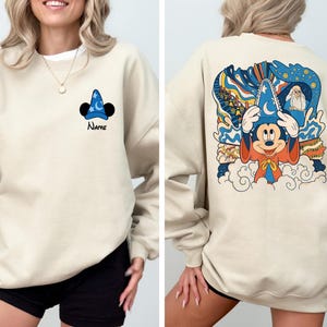 May include: A beige sweatshirt with a Mickey Mouse illustration wearing a sorcerer hat on the back. The front of the sweatshirt has a small Mickey Mouse ear and hat graphic with the word "Name" underneath.