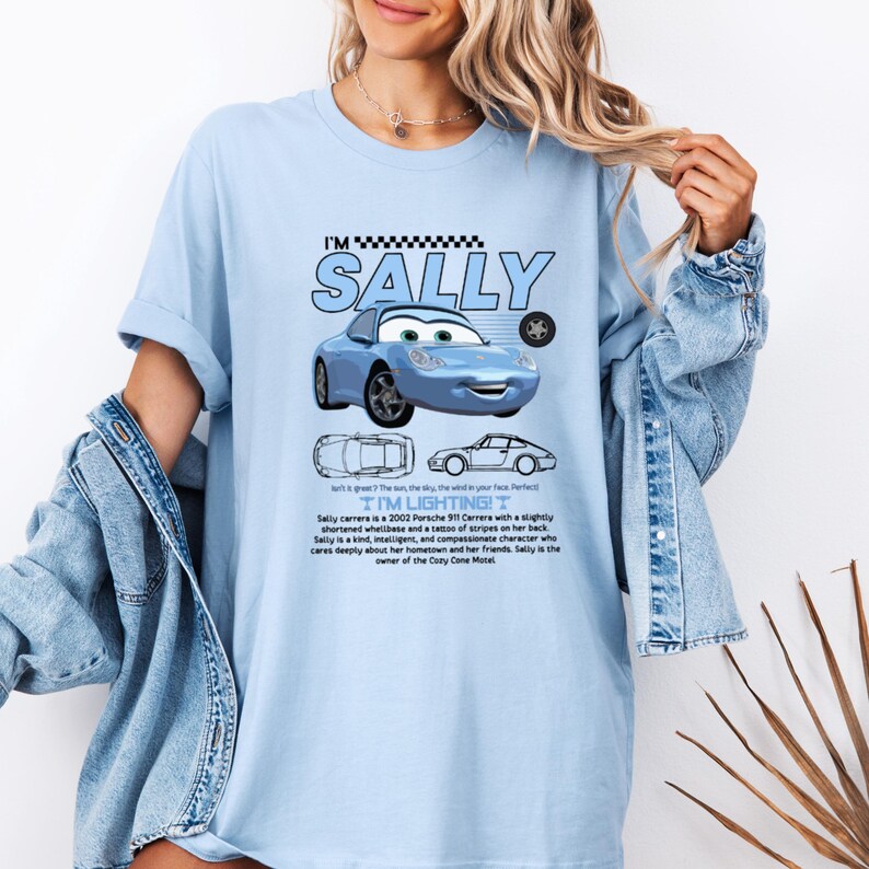 May include: Light blue t-shirt featuring a cartoon image of Sally Carrera, a blue Porsche 911. The shirt includes the text "I'M SALLY" and "I'M LIGHTING!" along with a description of the character. The shirt is a comfortable, casual style.