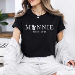 May include: Black t-shirt with the word "Minnie" in white, featuring a Minnie Mouse silhouette. Below the name is "Since 1928" in white. The shirt is short-sleeved.