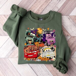 May include: Green sweatshirt with a Halloween-themed graphic featuring cartoon cars dressed up as monsters. The cars are surrounded by pumpkins and bats. The text "Boo!" is visible on the sweatshirt.