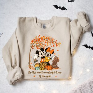 May include: Beige sweatshirt featuring Mickey and Minnie Mouse under a tree with orange leaves. The graphic includes pumpkins, a cowboy boot, and the text "It's the most wonderful time of the year."