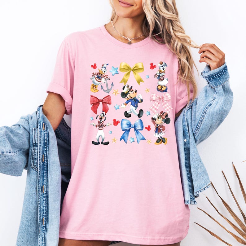 May include: Pink t-shirt with a colorful print of Mickey Mouse and friends wearing sailor outfits and holding bows. The print includes stars, anchors, and hearts.