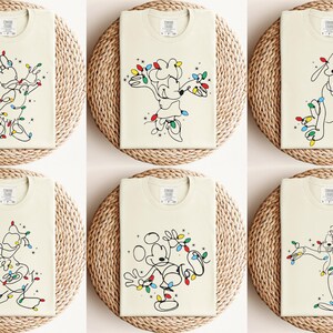 May include: Six cream-colored t-shirts with different cartoon character designs. Each shirt features a black line drawing of a character, adorned with colorful Christmas lights. The shirts are displayed on round, woven placemats.