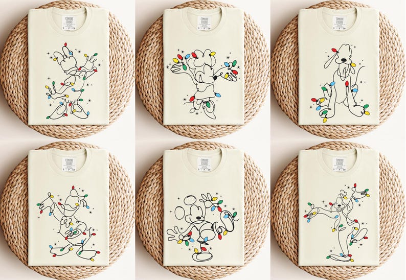 May include: Six cream-colored t-shirts with different cartoon character designs. Each shirt features a black line drawing of a character, adorned with colorful Christmas lights. The shirts are displayed on round, woven placemats.