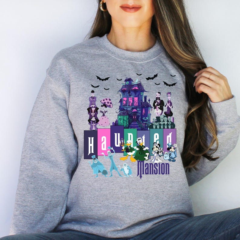 Puede incluir: Una sudadera gris con un estampado gr&aacute;fico de la atracci&oacute;n Haunted Mansion de Disneyland. El estampado presenta el edificio de la Haunted Mansion, fantasmas y personajes de la atracci&oacute;n. El texto "Haunted Mansion" est&aacute; impreso debajo del gr&aacute;fico.