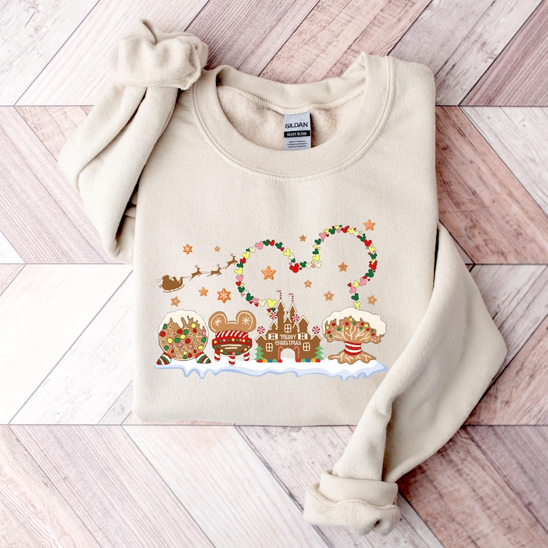 May include: A beige sweatshirt with a Christmas-themed design featuring a gingerbread castle, Mickey Mouse, and a Christmas tree. The design includes the text "Merry Christmas" and a garland of lights in the shape of Mickey Mouse ears.