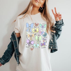 May include: White t-shirt with a colorful graphic of bows and the names of Disney princesses: Ariel,  Moana, Cinderella, Belle, Aurora, Merida, Tiana, Mulan, Rapunzel, Jasmine, Snow White.