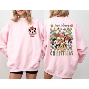 May include: A light pink sweatshirt featuring Minnie Mouse on the front with a red bow and the name "Name". The back of the sweatshirt has a graphic of Disney characters with the words "Very Merry CHRISTMAS".