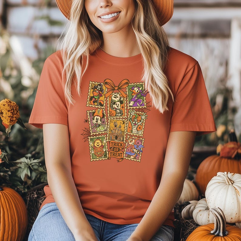 May include: Orange t-shirt featuring a collection of Halloween-themed postage stamp designs. The stamps showcase various cartoon characters and the phrase "Trick or Treat". A large orange bow is above the stamps.