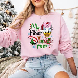 May include: Pink sweatshirt with a Christmas-themed Disney graphic featuring a cartoon duck wearing a Santa hat and the text "My First Disney Trip".