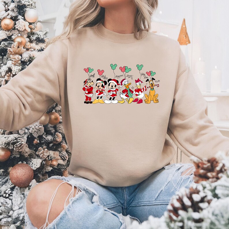 May include: Beige sweatshirt featuring a festive graphic of Disney characters dressed in holiday attire. Mickey, Minnie, Donald, Daisy, Goofy, and Pluto are depicted holding balloons. The design includes red, green, and white colors, perfect for the holiday season.