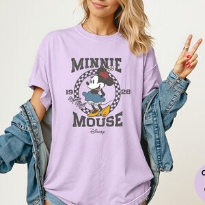May include: A light purple t-shirt featuring a distressed graphic of Minnie Mouse. The design includes the text "MINNIE MOUSE" and the years "1928". Minnie Mouse is depicted in her classic outfit. The shirt is a Comfort Colors Orchid.