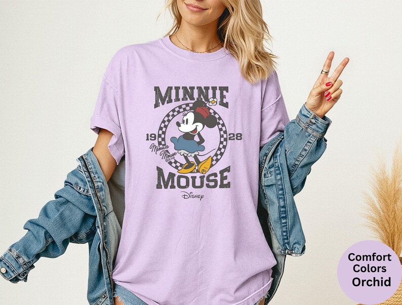 May include: A light purple t-shirt featuring a distressed graphic of Minnie Mouse. The design includes the text "MINNIE MOUSE" and the years "1928". Minnie Mouse is depicted in her classic outfit. The shirt is a Comfort Colors Orchid.
