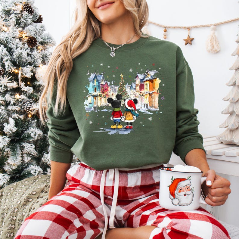 Op de afbeelding: Groen sweatshirt met ronde hals met een winterse sc&egrave;ne met Mickey en Minnie Mouse. De afbeelding toont het paar voor een besneeuwd dorp met een kerstboom. De persoon draagt een rood-witte geruite pyjamabroek en houdt een kerstmanmok vast.